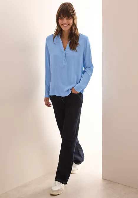 Frau mit hellblauer Bluse, dunkler Hose und weißen Sneakers steht lächelnd im Flur.
